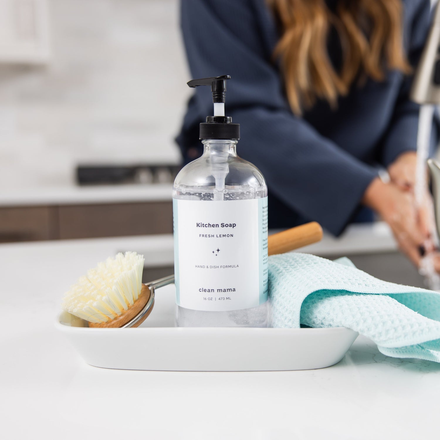 Bottle of kitchen soap on a kitchen counter with a person washing dishes in the background.